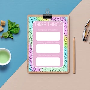 Peut inclure: Un presse-papiers rose et arc-en-ciel avec le texte "THREE THINGS" et trois lignes vides pour écrire. Le texte sous les lignes indique "Je suis fier de moi pour", "Quelque chose de bien que j'ai fait aujourd'hui", et "Je prévois d'accomplir demain".