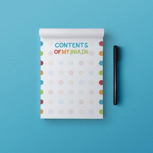 May include: A white notepad with a colourful polka dot pattern and the text "CONTENTS OF MY BRAIN:" on the front. A black pen is lying next to the notepad.