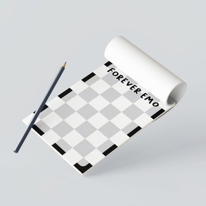 Puede incluir: Un bloc de notas blanco con un patrón de cuadros en blanco y negro y el texto "FOREVER EMO" en la portada. Un lápiz negro está acostado junto al bloc de notas.