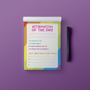 Op de afbeelding: Een notitieboekje in regenboogkleuren met de tekst "Affirmation van de dag" bovenaan. Het notitieboekje heeft lijnen om affirmaties, dagelijkse trainingsschema's en doelen voor zelfzorg op te schrijven.