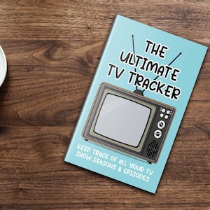 Könnte beinhalten: Ein blaues Buch mit dem Titel "THE ULTIMATE TV TRACKER" in weißer Schrift. Auf dem Cover befindet sich ein Cartoon-Bild eines alten Fernsehgeräts. Der Text unter dem Bild lautet "KEEP TRACK OF ALL YOUR TV SHOW SEASONS & EPISODES".