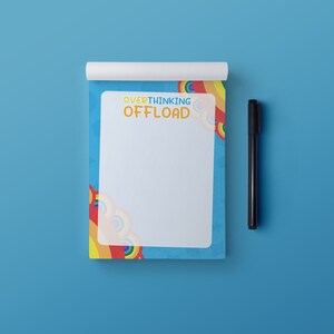 Peut inclure: Un bloc-notes bleu avec un motif arc-en-ciel et le texte "Overthinking Offload" sur le devant. Un stylo noir est posé à côté du bloc-notes.