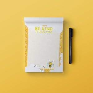 May include: A white notepad with a yellow honeycomb pattern border and the text "BE KIND TO YOUR MIND". A cartoon bee is flying near the bottom of the notepad. A black pen is lying on a yellow surface next to the notepad.