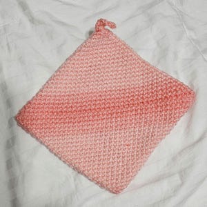 May include: A pink knitted pot holder with a loop handle. The pot holder is made with a gradient yarn that transitions from a lighter pink to a darker pink.