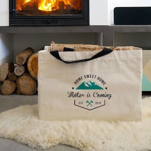 Peut inclure: Un sac fourre-tout en toile crème avec des poignées noires, imprimé avec "HOME SWEET HOME Winter is Coming EST. 2010" en teal et noir. Le sac est rempli de bois de chauffage et posé sur un tapis blanc moelleux près d'une cheminée.