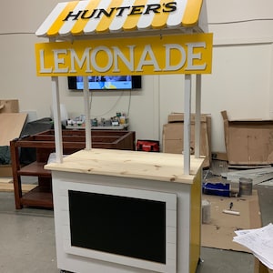 May include: A wooden lemonade stand with a yellow and white striped awning. The sign reads "HUNTER'S LEMONADE" in black and white letters. The stand has a chalkboard and is painted white and yellow.