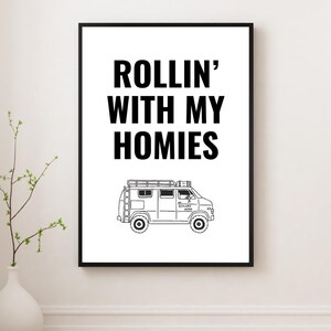 May include: Black framed print with the text "ROLLIN' WITH MY HOMIES" above a line drawing of a van. The van has a roof rack and the words "EXPLORE MORE" on the side. The print is displayed on a white wall.