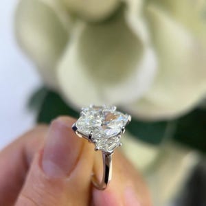 May include: A pear-shaped diamond engagement ring with a three-stone setting. The center stone is a large, brilliant cut diamond, flanked by two smaller, baguette-cut diamonds.
