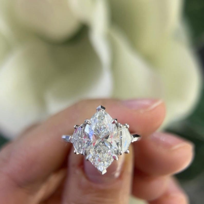 May include: A three-stone engagement ring with a large, central, marquise-shaped diamond and two smaller, pear-shaped diamonds on either side. The ring is set in a white gold band.