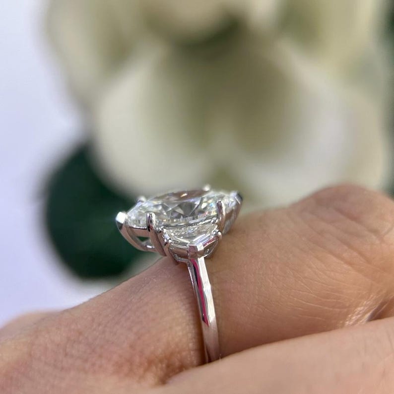 May include: A silver ring with a large, clear, oval-shaped center stone and two smaller, clear, triangular stones on either side.