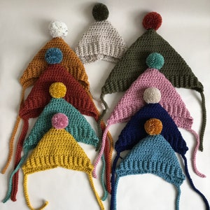 Peut inclure: Plusieurs bonnets crochetés faits à la main de différentes couleurs, dont jaune moutarde, sarcelle et bleu marine. Chaque bonnet est orné d'un pompon et de liens. Les bonnets sont disposés sur une surface blanche.