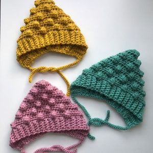 May include: Three handmade crochet baby bonnets in mustard yellow, dusty rose, and teal. Each bonnet features a textured, bobble stitch design and ties under the chin. The bonnets are displayed on a white surface.