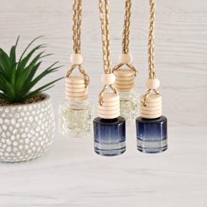 May include: Four small glass car air freshener bottles hanging from braided gold and white cords. Two bottles are clear with a faceted texture, and two are a deep navy blue gradient. Each bottle features a light-colored wooden screw-top cap.
