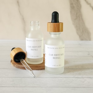 May include: Two frosted glass bottles with bamboo lids and black droppers. The bottles are labeled "The Bath and Wick Shop" and "Car Diffuser Refill" with a 1 oz (30 ml) size.