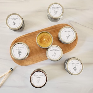 May include: Seven round metal tins of beeswax candles with white labels featuring botanical illustrations and scent names including Rosemary Sage, Palo Santo Patchouli, Lavender, Teakwood Mahogany, Vanilla Brown Sugar, Sandalwood Vanilla, and Eucalyptus Mint.