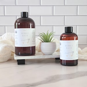 May include: Two brown bottles of bubble bath with black caps, labeled "The Bath and Milk Shop." A small white pot with a green succulent sits between the bottles on a white and black tray. The background is a white tile wall.