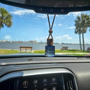 May include: A blue glass car air freshener hanging from a rearview mirror. The air freshener has a wooden top and a brown string. The background shows a sunny day with a body of water and palm trees.