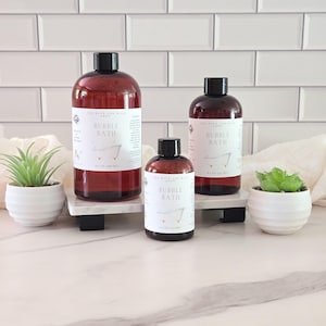 May include: Three amber plastic bottles of bubble bath with black caps and white labels featuring the text BUBBLE BATH and a small bathtub illustration. The bottles are in three sizes, labeled 16 fl oz, 8 fl oz, and 4 fl oz, arranged on white stands.