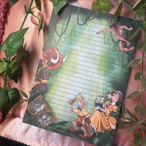 Op de afbeelding: Gelijnd notitieblok met een junglescène met stripfiguren. Het ontwerp bevat Tarzan, Jane en andere personages. Het notitieblok heeft een groen-bruine achtergrond met een roze rand en is omgeven door groene bladeren.