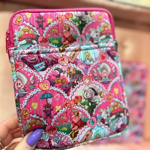 Könnte beinhalten: Rosa Reißverschlusstasche mit einem farbenfrohen Alice im Wunderland-Muster. Das Design umfasst Charaktere wie die Grinsekatze und den Hutmacher, mit Herzen und anderen thematischen Elementen. Die Tasche hat einen rosa Reißverschluss und eine rechteckige Form.