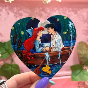 May include: A heart-shaped illustration depicts a romantic scene with a red-haired woman and a man in a boat under a full moon. The artwork features a blue and green color palette, with details of water, reeds, and a yellow fish.