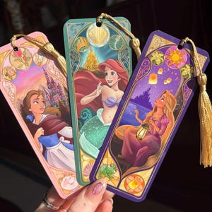 Puede incluir: Tres marcapáginas coloridos con ilustraciones de las princesas Disney Bella, Ariel y Rapunzel. Cada marcapáginas tiene una borla dorada y un borde decorativo. El marcapáginas de Bella es rosa, el de Ariel es turquesa y el de Rapunzel es morado.