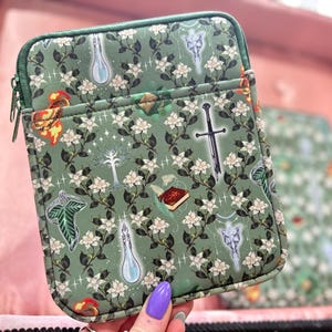 May include: Green zippered pouch featuring a pattern of white flowers and fantasy symbols. The pouch has a green zip and trim. The background is a light pink.