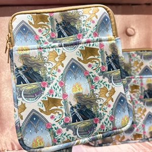 May include: Two zippered pouches featuring a repeating pattern of a woman with a sword, dragons, and gothic windows. The background is cream with pink roses and green leaves. The zipper is gold.