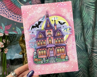 May include: A Halloween art print depicting a haunted house in pink and purple hues, with bats, pumpkins, and a full moon. The illustration has a whimsical style with a pink speckled background, perfect for Halloween decorations.