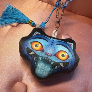 May include: A blue cat-shaped keychain with a cartoonish design. The cat has large orange eyes, sharp teeth, and a tongue that reads "HELLO FRIEND". It is attached to a blue tassel and a silver clasp.