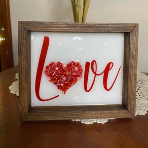 Love glass and resin art.  This art is 5 inches tall and 6 inches wide.  The O in love is formed by a heart shape filled with glass and beads and then a beautiful layer of resin is added.
