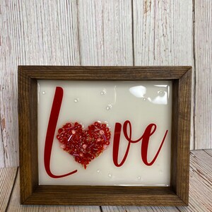 May include: A framed piece of art with the word "LOVE" written in red cursive. A red heart made of beads is in the center of the word.