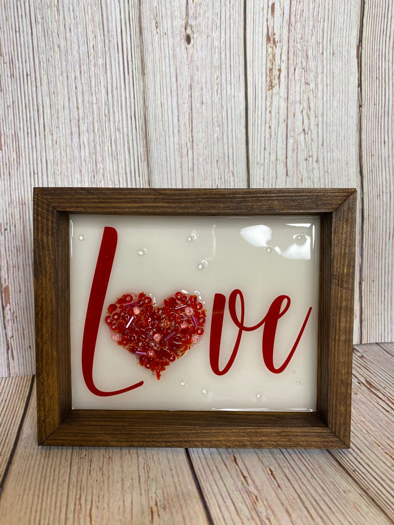 May include: A framed piece of art with the word "LOVE" written in red cursive. A red heart made of beads is in the center of the word.