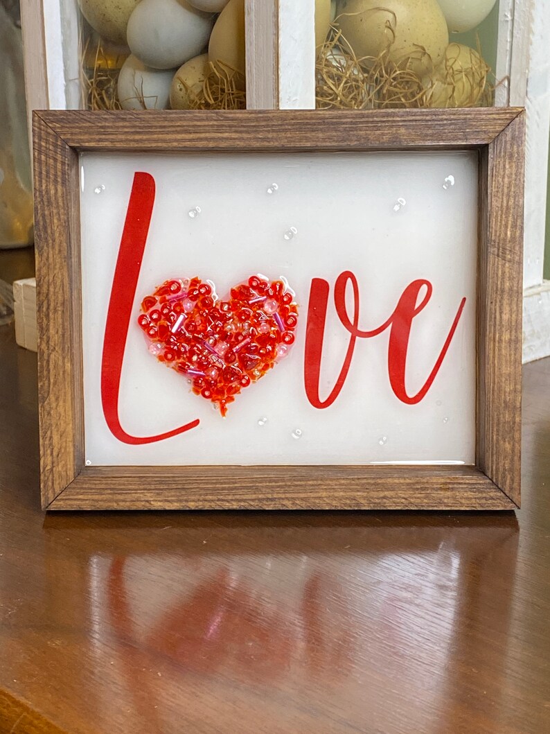 May include: A framed sign with the word "LOVE" in red lettering. A red heart made of beads is in the center of the word.
