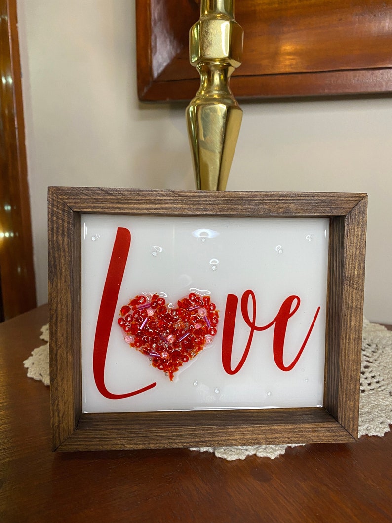 Love glass and resin art.  This art is 5 inches tall and 6 inches wide.  The O in love is formed by a heart shape filled with glass and beads and then a beautiful layer of resin is added.