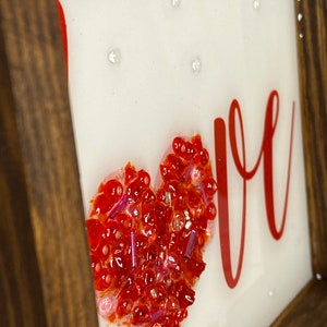 May include: A framed piece of art with the word "LOVE" in red lettering. A red heart made of small red and pink beads is in the center of the frame.