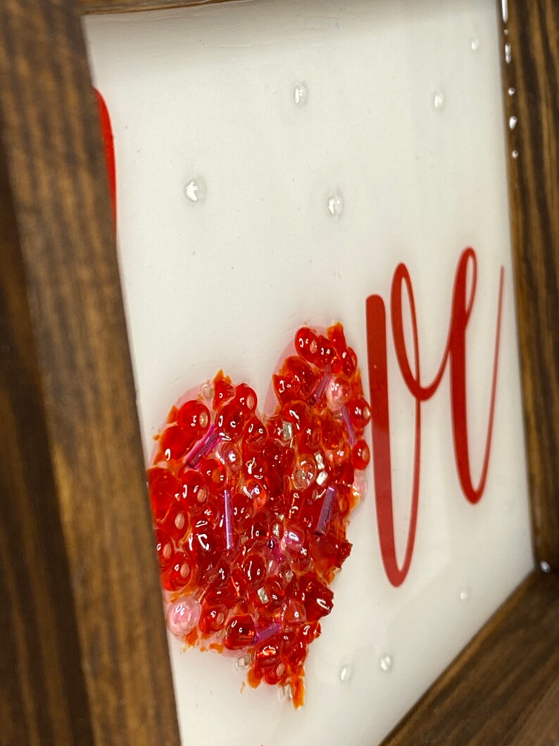 May include: A framed piece of art with the word "LOVE" in red lettering. A red heart made of small red and pink beads is in the center of the frame.