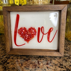 May include: A framed sign with the word "LOVE" in red lettering. A heart shape made of red and pink beads is in the center of the sign.