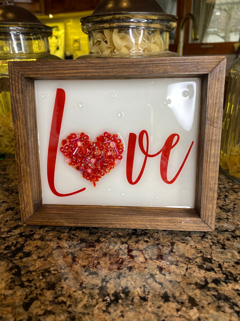 May include: A framed sign with the word "LOVE" in red lettering. A heart shape made of red and pink beads is in the center of the sign.