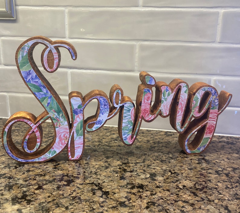 以下が含まれることがあります： 花柄の「Spring」という文字が書かれた木製看板。文字は茶色で、ピンク、青、緑の花柄のデザインが施されています。