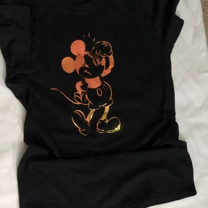 以下が含まれることがあります： 黒いTシャツ。金とオレンジ色の漫画のネズミのグラフィックが特徴です。ネズミはクラシックなポーズで、腕を上げています。デザインはシャツの前面の中央に配置されています。