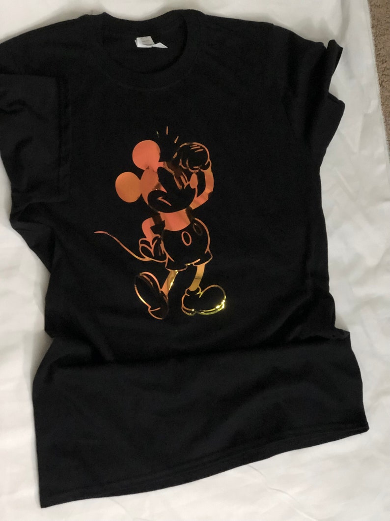 以下が含まれることがあります： 黒いTシャツ。金とオレンジ色の漫画のネズミのグラフィックが特徴です。ネズミはクラシックなポーズで、腕を上げています。デザインはシャツの前面の中央に配置されています。
