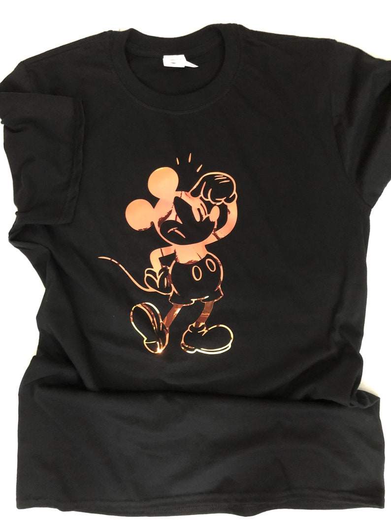 以下が含まれることがあります： 黒いTシャツに、遊び心のあるポーズのミッキーマウスの銅色のグラフィックが描かれています。 このデザインは、象徴的な漫画のキャラクターの様式化されたイラストです。 シャツは柔らかい素材でできており、クルーネックです。