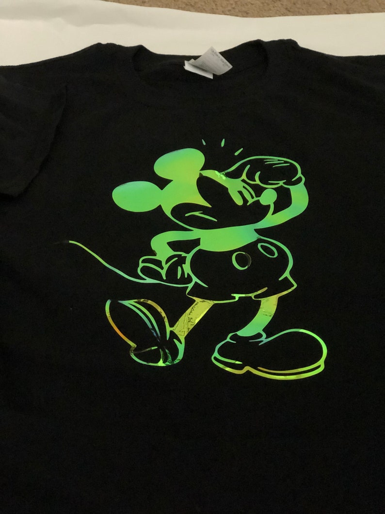 以下が含まれることがあります： 黒いTシャツ。緑と黄色のグラデーションでミッキーマウスが描かれています。漫画のキャラクターは走っているポーズで描かれています。Tシャツはクルーネックです。