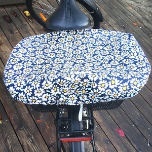 Könnte beinhalten: Ein schwarzes Fahrrad mit einem weißen Korbüberzug mit einem Gänseblümchenmuster. Das Fahrrad hat einen schwarzen Sattel und einen schwarzen hinteren Kotflügel mit einem roten Reflektor.