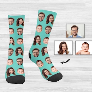 May include: A pair of turquoise blue socks with a repeating pattern of different faces. The faces include a man, a woman, and a baby. The socks are shown on a light wood background.