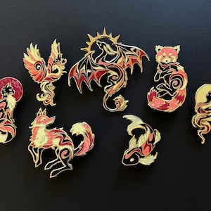 May include: A collection of seven gold-toned metal enamel pins featuring stylised animal and mythical creature designs. The pins include a cat, a phoenix, a dragon, a red panda, a fox, a koi fish, and a snake, all with black, red, and cream accents.