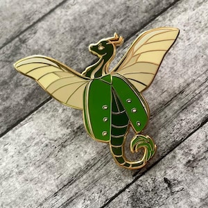 May include: A gold-coloured enamel pin of a stylised dragon-like creature with green and tan wings and a green jacket. The pin is set against a grey wooden background.