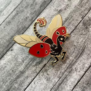 May include: A gold-colored metal pin featuring a stylized creature with a scorpion tail and moth wings. The wings are cream and red with black spots. The creature's body is black with gold accents.