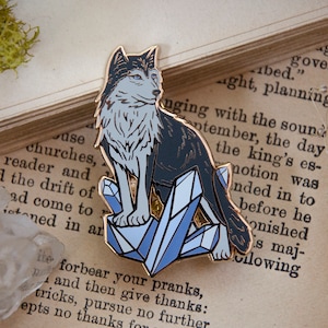 May include: A black and white enamel pin of a wolf sitting on a bed of blue crystals. The wolf has a white chest and a blue eye. The pin is on a book with text.